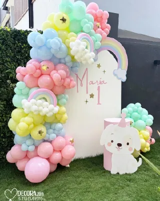 Mampara blanca con arcoíris de cartón y guirnalda de globos pastel sobre pasto